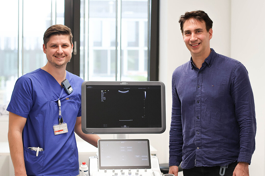 In einem hellen Raum stehen zwei Herren neben einem Ultraschallgerät. Einer der beiden trägt einen blauen Arztkittel und legt seine Hand auf dem Gerät ab. Der andere trägt ein dunkelblaues Leinenhemd. Beide lächeln in die Kamera