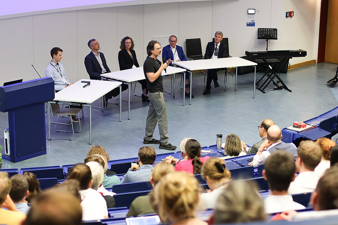 Fünf Personen sitzen an einer Tischreihe auf einer Tribüne im Hörsaal. Auf den Plätzen in den ersten Reihen des Hörsaals sitzen zuhörende Personen. Vor der Sitzreihe steht ein Mann mit Mikrofon in der Hand.