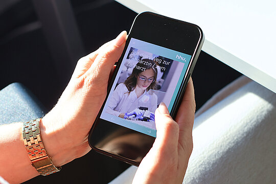Eine Frau hält ein Handy in der Hand. Darauf ist ein Bild zu sehen auf dem eine junge Frau in weißem Kittel an einem Zahnmodell bohrt. Auf dem Bild steht der Titel "Auf dem Weg zur Zahnärztin / zum Zahnarzt".