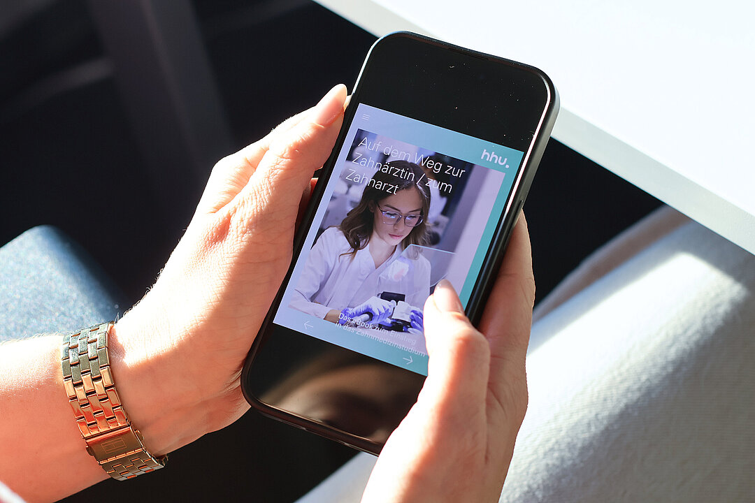 Eine Frau hält ein Handy in der Hand. Darauf ist ein Bild zu sehen auf dem eine junge Frau in weißem Kittel an einem Zahnmodell bohrt. Auf dem Bild steht der Titel "Auf dem Weg zur Zahnärztin / zum Zahnarzt".