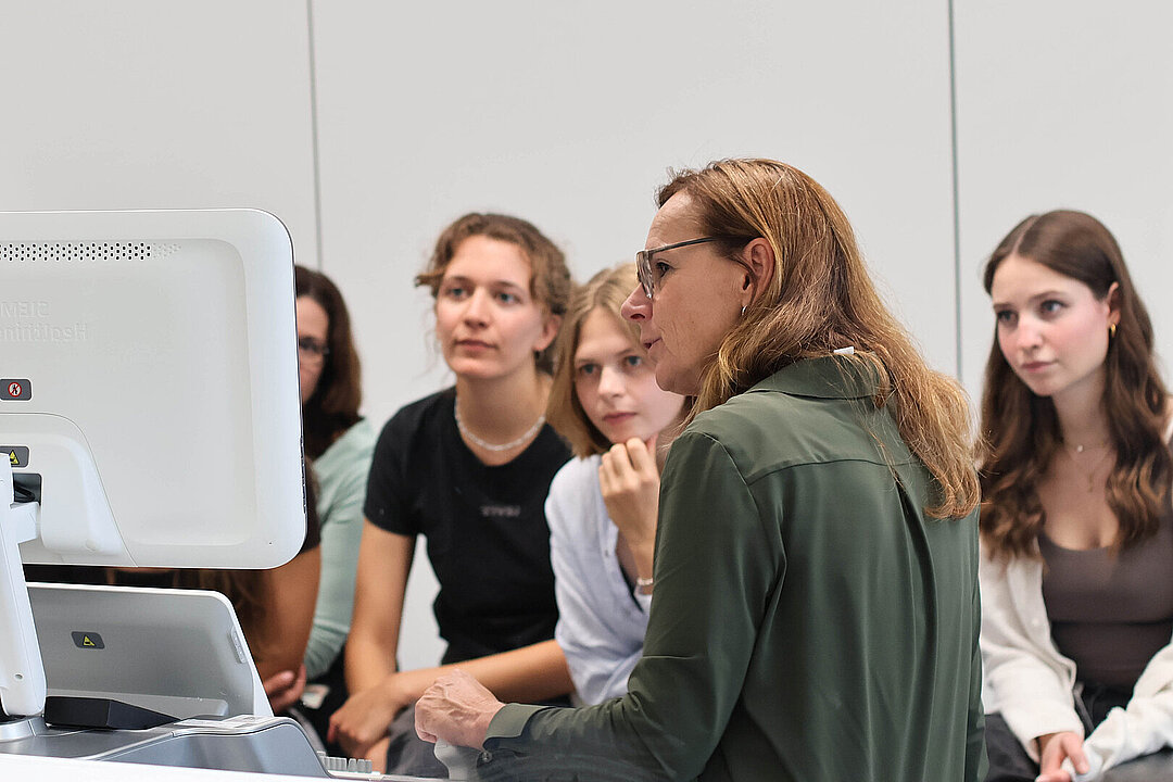 Eine Frau mit Olivgrünem Oberteil blickt auf ein Ultraschallgerät und scheint für jungen Studentinnen, welche neben ihr sitzen, das Gerät zu erklären.