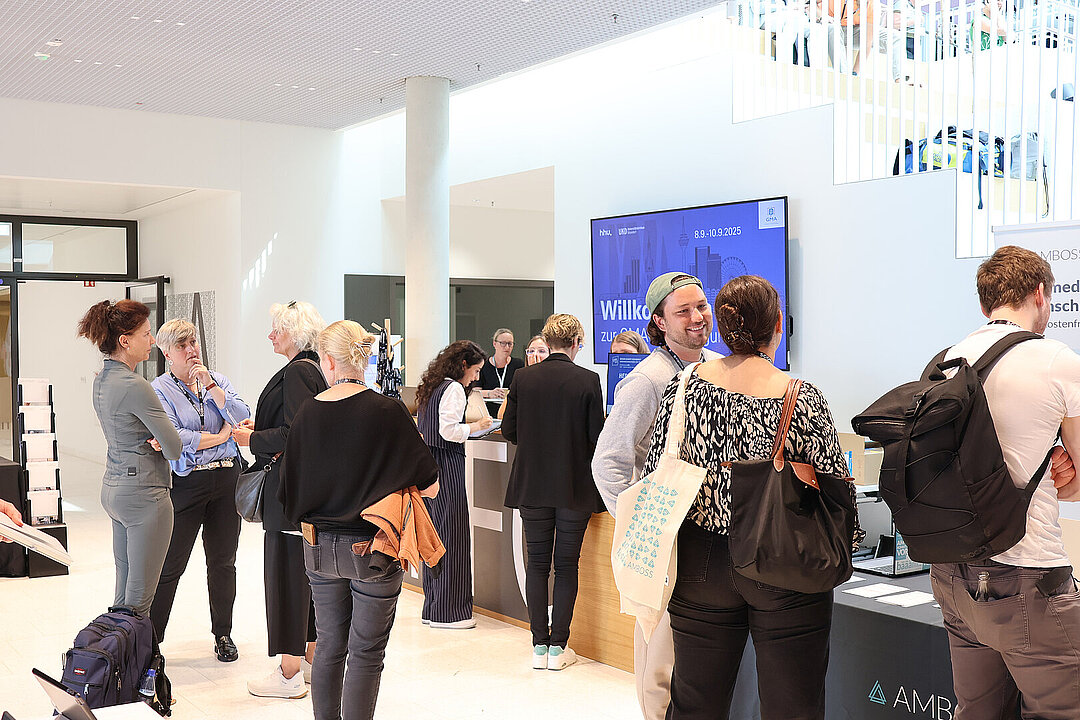 Mehrere Personen sammeln sich im Foyer eines hellen, modernen Gebäudes. Sie stehen an der Anmeldung an und unterhalten sich lächelnd miteinander.