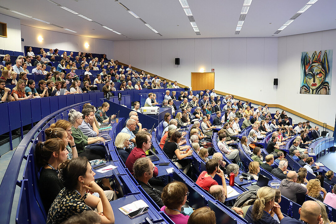 Blick in einen Hörsaal. Die Sitzreihen sind fast vollständig von Erwachsenen besetzt und alle schauen nach vorne. Die Sitzreihen haben dunkelblaue Stühle und an der Wand des Hörsaals hängt ein abstraktes Kunstwerk.