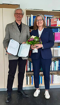 links Professor Frank Leinen mit Abschiedsurkunde und rechts Dekanin Ulli Seegers mit einem Blumenstrauß stehen vor einem Bücherregal.