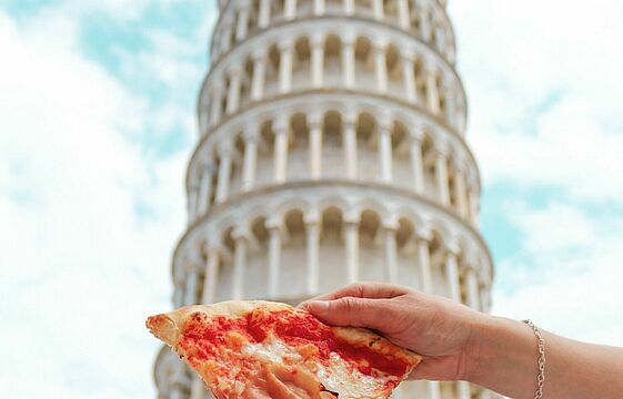 Frauenhand mit Pizzaecke vor dem Schiefen Turm von Pisa