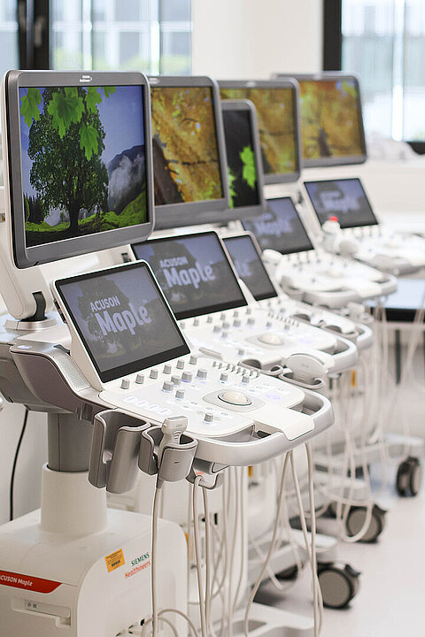 Auf dem Bild sind fünf hochmoderne Ultraschallgeräte zu sehen, die in einem Raum hintereinander gereiht aufgestellt sind. Der Bildschirm zeigt einen Bildschirmschoner. 