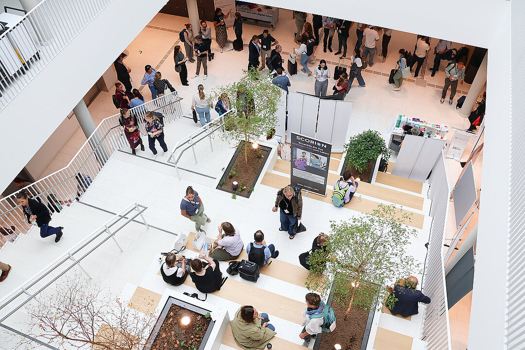 Blick von oben in ein Foyer eines hellen, modernen Gebäudes. Menschen halten sich im Foyer auf und sitzen auf Treppen