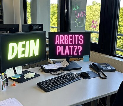 Ein Schreibtisch in einem Büro mit Ausblick ins Grüne, auf den Bildschirmen steht: Dein Arbeitsplatz?