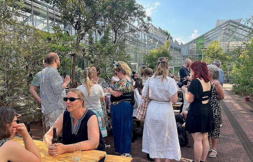Menschen beim Sommerfest sitzen an Biertischen und stehen vor den Gewächshäusern