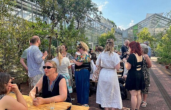 Menschen beim Sommerfest sitzen an Biertischen und stehen vor den Gewächshäusern
