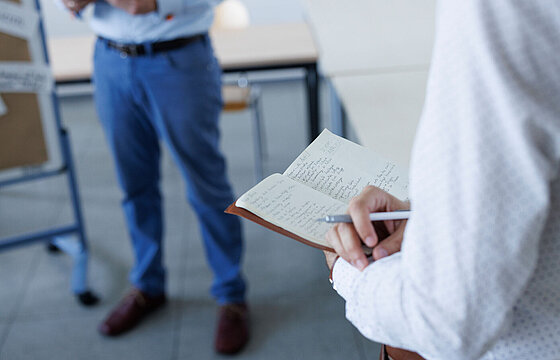 Eine Person mit Notizbuch und die Beine einer anderen Person sind zu sehen