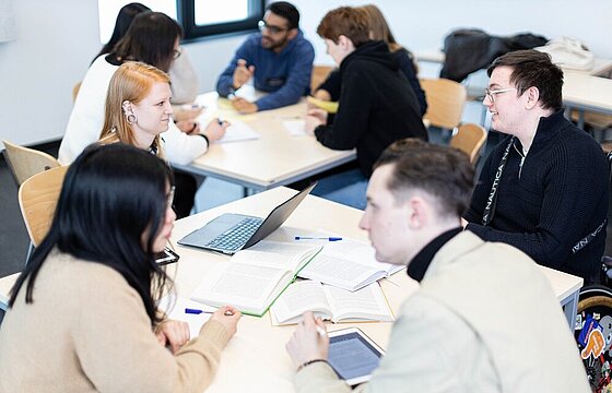 Studierende sitzen in einem Seminarraum in Gruppen an kleinen Tischen.
