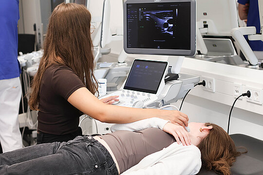 Eine junge Frau bedient ein Ultraschallgerät und schallt eine weitere junge Frau am Hals, welche auf einer Liege liegt. Beide schauen in Richtung des Ultraschall-Bildschirms